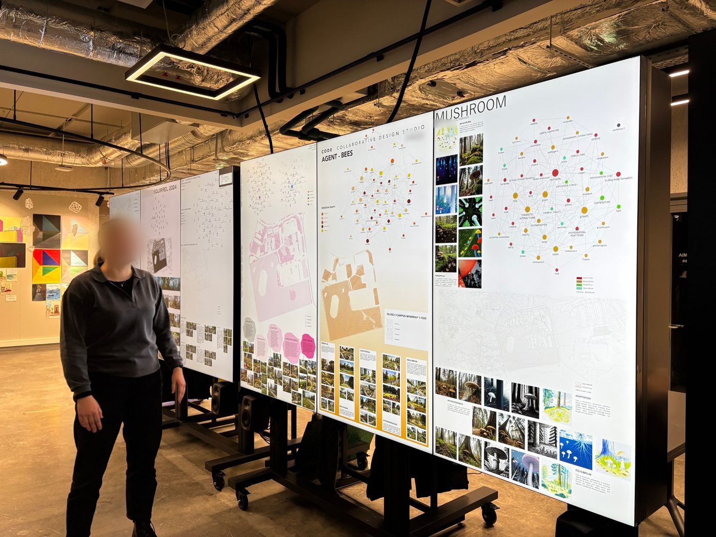 A wall of tall screens, each showing one student&rsquo;s poster for an agent of the campus; the bees, the mushroom and the squirrel are in view. Each poster has a graph of linked notes, a map of the campus and rows of generated images. A person stands at the left, looking at the screens; the face is blurred.
